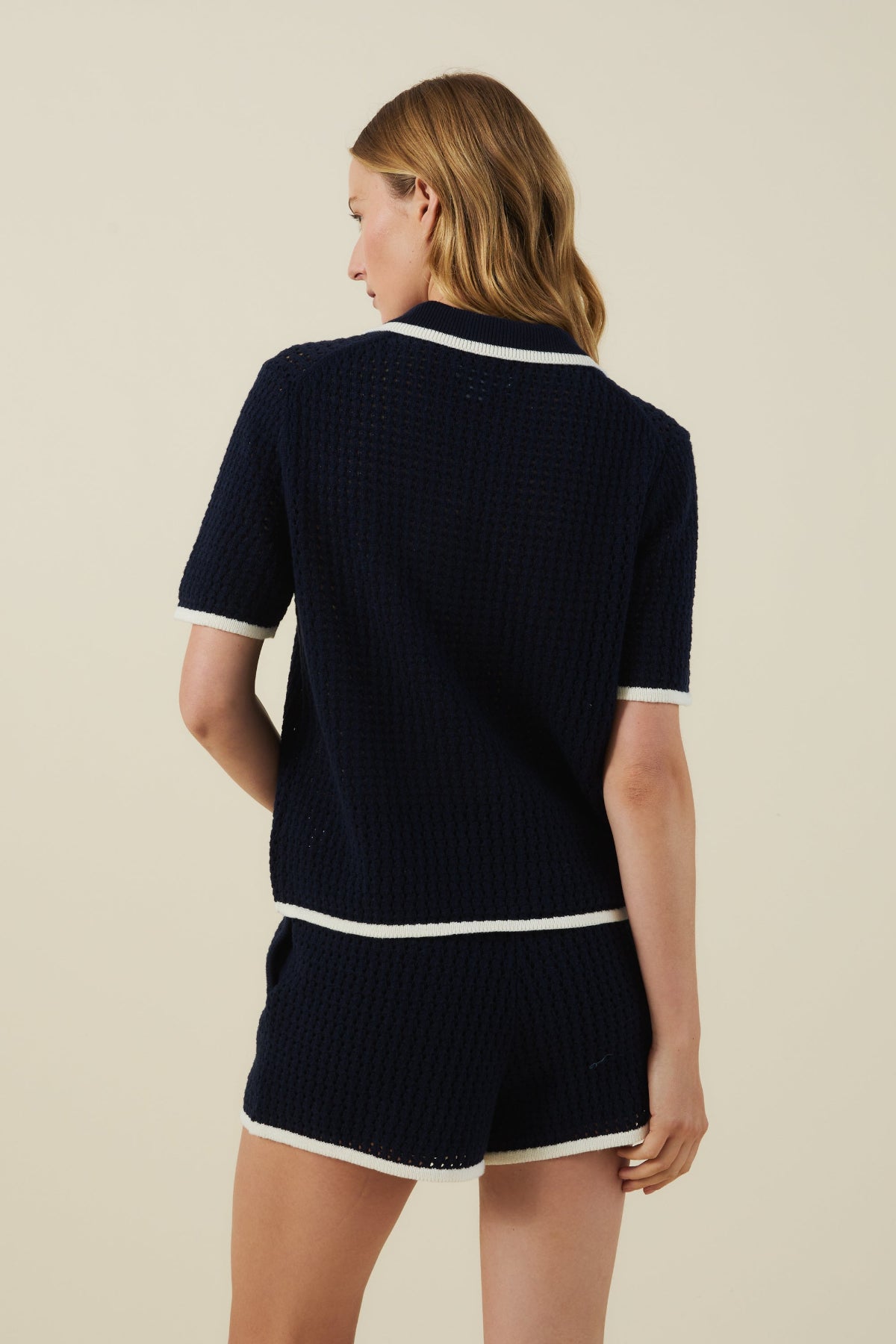 A woman stands facing away, wearing the Velvet by Graham & Spencer LUMAE SWEATER POLO—a sporty-chic navy short-sleeve top—paired with matching shorts featuring white trim, set against a plain beige background.-41460579008705