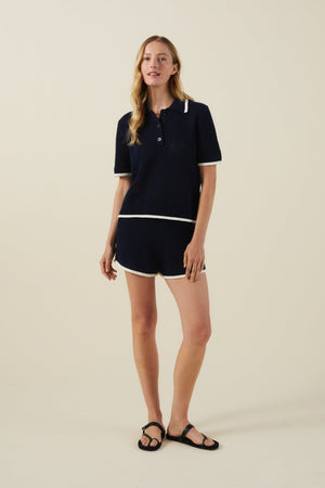 A woman wears the LUMAE SWEATER POLO by Velvet by Graham & Spencer— a sporty-chic navy short-sleeve knit polo shirt with white trim, 100% cotton—paired with matching shorts and black sandals against a beige background.