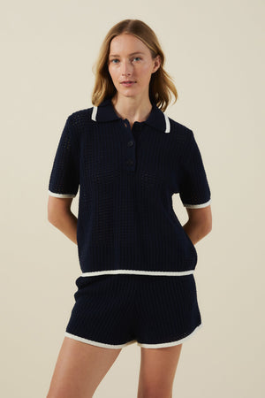 A woman stands against a plain background wearing the LUMAE SWEATER POLO by Velvet by Graham & Spencer—a sporty-chic navy short-sleeve knit polo shirt paired with matching shorts featuring white trim.