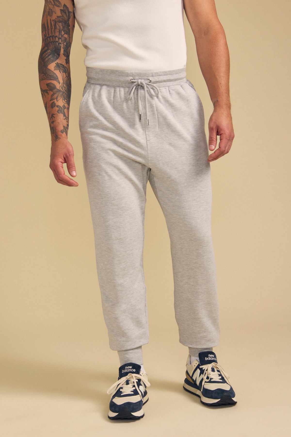 A person wears the JACOBY JOGGER by Velvet by Graham & Spencer—a light gray drawstring sweatpant—paired with a white t-shirt, blue and white sneakers, and light gray socks, standing against a beige background.-41702026379457