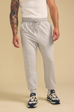 A person wears the JACOBY JOGGER by Velvet by Graham & Spencer—a light gray drawstring sweatpant—paired with a white t-shirt, blue and white sneakers, and light gray socks, standing against a beige background.