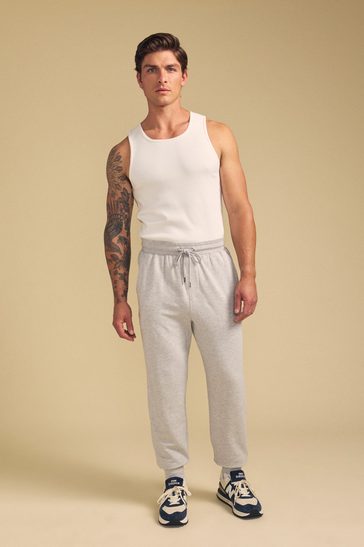 A man in the JACOBY JOGGER by Velvet by Graham & Spencer, a white tank top, and navy sneakers stands against a beige background, showcasing a large tattoo on one arm.-41702026346689