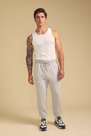A man in the JACOBY JOGGER by Velvet by Graham & Spencer, a white tank top, and navy sneakers stands against a beige background, showcasing a large tattoo on one arm.