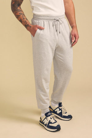 A person wearing a white shirt, Velvet by Graham & Spencer’s JACOBY JOGGER in light gray, and navy blue New Balance sneakers stands with one hand in their pocket against a beige background.
