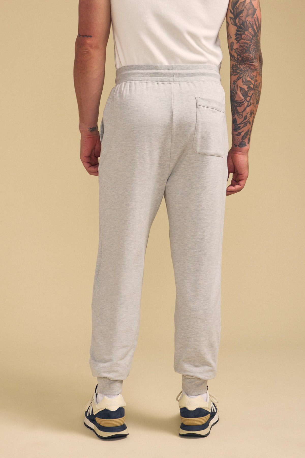 A person wearing the JACOBY JOGGER by Velvet by Graham & Spencer in light gray, paired with a white T-shirt and sneakers, stands with their back to the camera against a beige background.