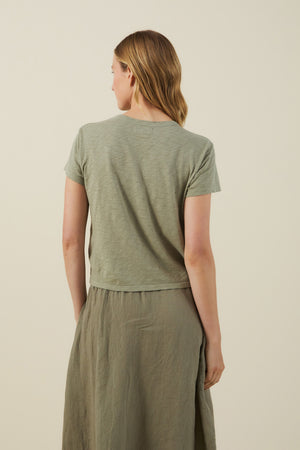 Back view of a woman wearing the Velvet by Graham & Spencer MARIKA TEE in green, made from soft cotton slub with a relaxed boxy fit, paired with a matching green skirt.