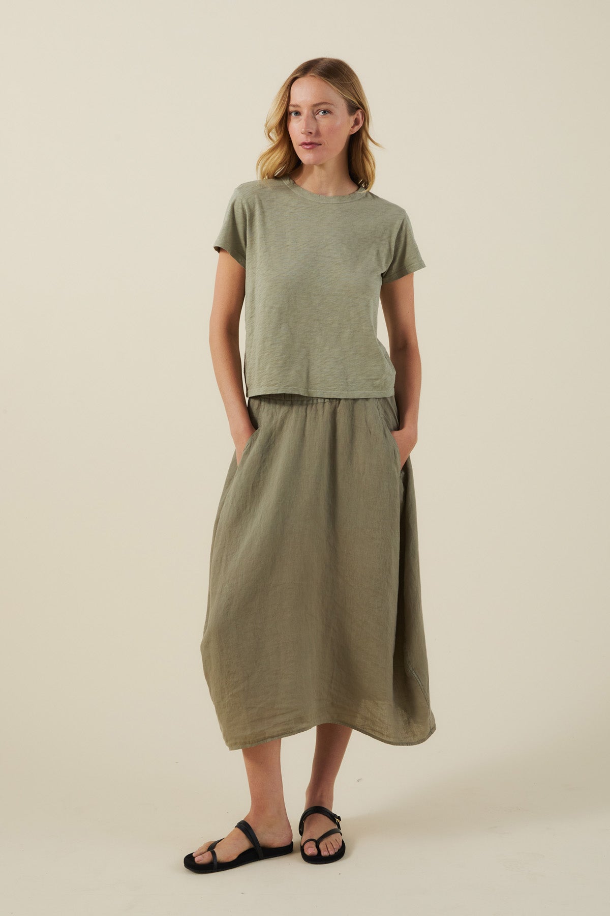 A woman stands with hands in pockets, wearing a light green tee, the FAE LINEN SKIRT by Velvet by Graham & Spencer—an olive A-line linen skirt—and black sandals, against a plain light background.-41645029064897