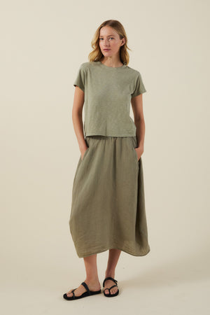A woman stands with hands in pockets, wearing a light green tee, the FAE LINEN SKIRT by Velvet by Graham & Spencer—an olive A-line linen skirt—and black sandals, against a plain light background.