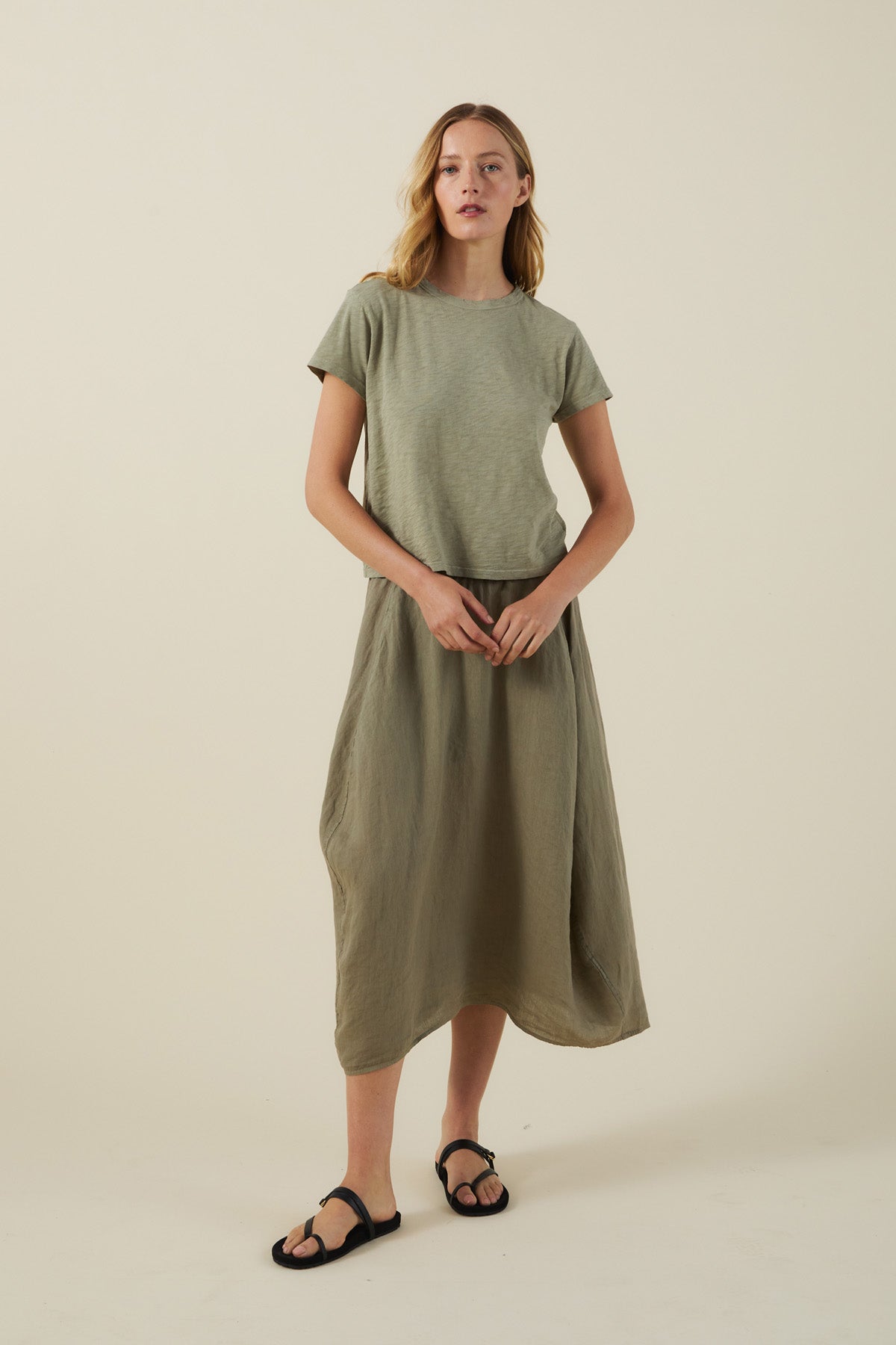A woman stands against a plain background wearing a light green t-shirt, the FAE LINEN SKIRT by Velvet by Graham & Spencer in olive green with an A-line silhouette, and black sandals.-41645029195969