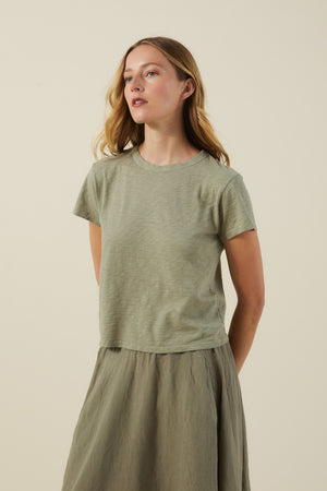 A woman with light brown hair wears the MARIKA TEE by Velvet by Graham & Spencer, a short-sleeved, light olive green cotton slub shirt with a relaxed boxy fit, paired with a matching skirt against a plain beige background.