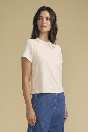 A person with long brown hair wears a short-sleeve beige MARIKA TEE by Velvet by Graham & Spencer, crafted from soft cotton slub in a relaxed boxy fit, paired with blue jeans. They stand against a plain beige background, looking slightly to the side.