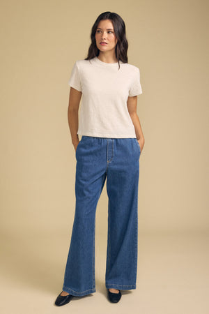 A person stands against a beige background, wearing the MARIKA TEE by Velvet by Graham & Spencer—a short-sleeve beige top made from cotton slub in a relaxed boxy fit—paired with wide-leg blue jeans and black shoes, hands in their pockets.