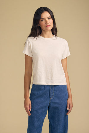 A woman with long brown hair wears a plain beige MARIKA TEE by Velvet by Graham & Spencer in a relaxed boxy fit with blue jeans, standing against a tan background.
