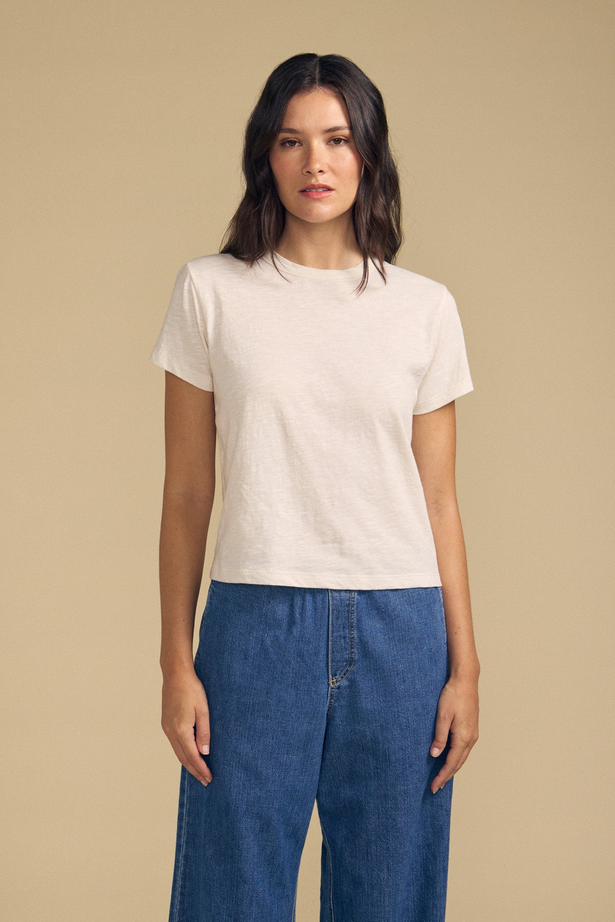 A woman stands against a beige background, wearing the MARIKA TEE by Velvet by Graham & Spencer in a relaxed boxy fit crafted from soft cotton slub, paired with blue jeans. She faces forward with a neutral expression.