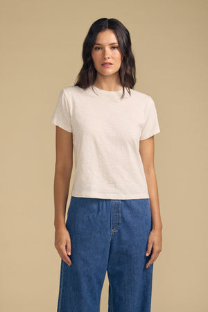 A woman stands against a beige background, wearing the MARIKA TEE by Velvet by Graham & Spencer in a relaxed boxy fit crafted from soft cotton slub, paired with blue jeans. She faces forward with a neutral expression.