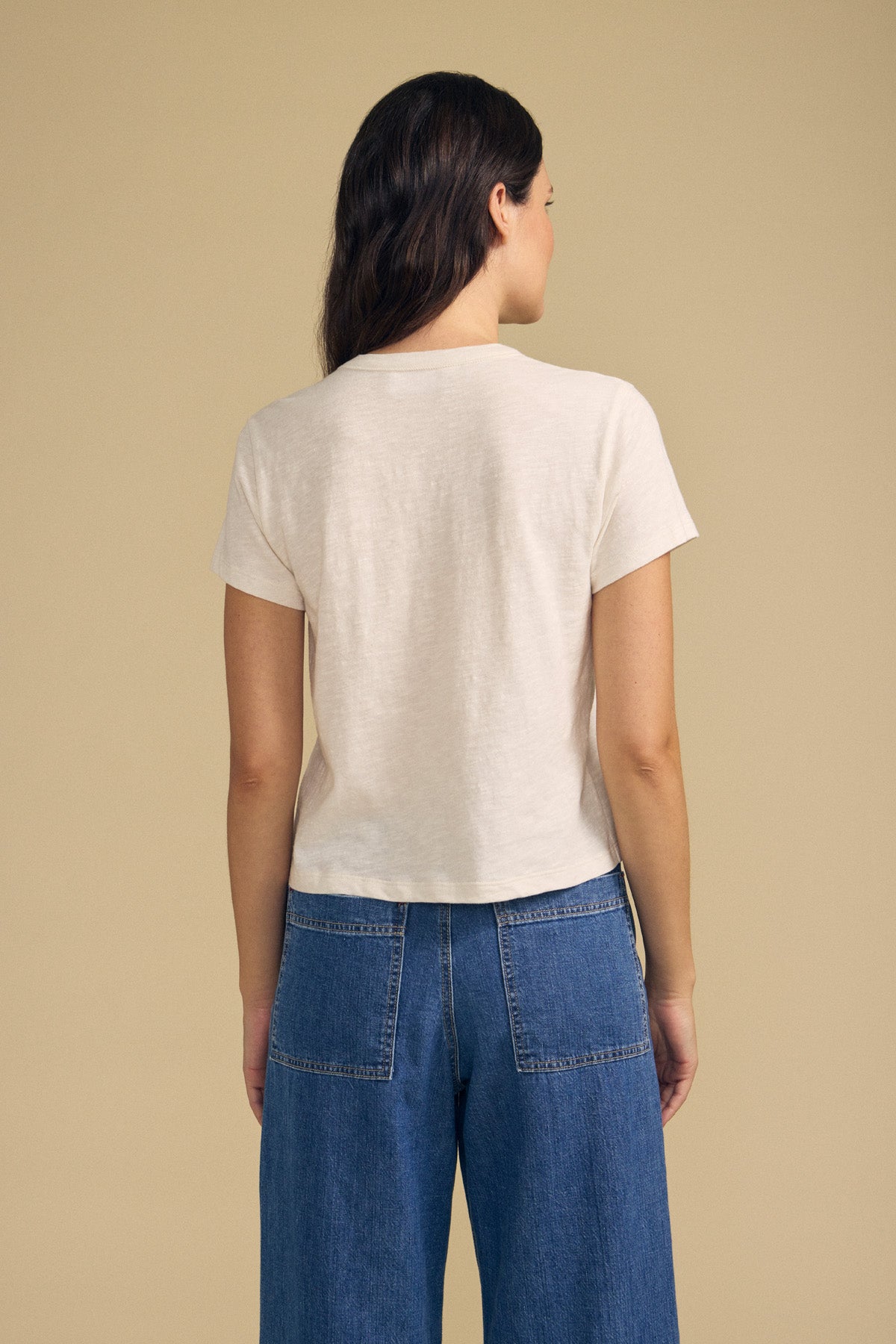 Person standing with back to camera, wearing a short-sleeve off-white MARIKA TEE by Velvet by Graham & Spencer in a relaxed boxy fit and high-waisted blue jeans, crafted from soft cotton slub, against a plain beige background.-41844137296065