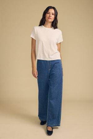 A woman stands against a beige background, wearing a plain white t-shirt, the SYD JEAN by Velvet by Graham & Spencer—relaxed fit wide-leg denim jeans with an elastic drawstring waist—and black shoes.