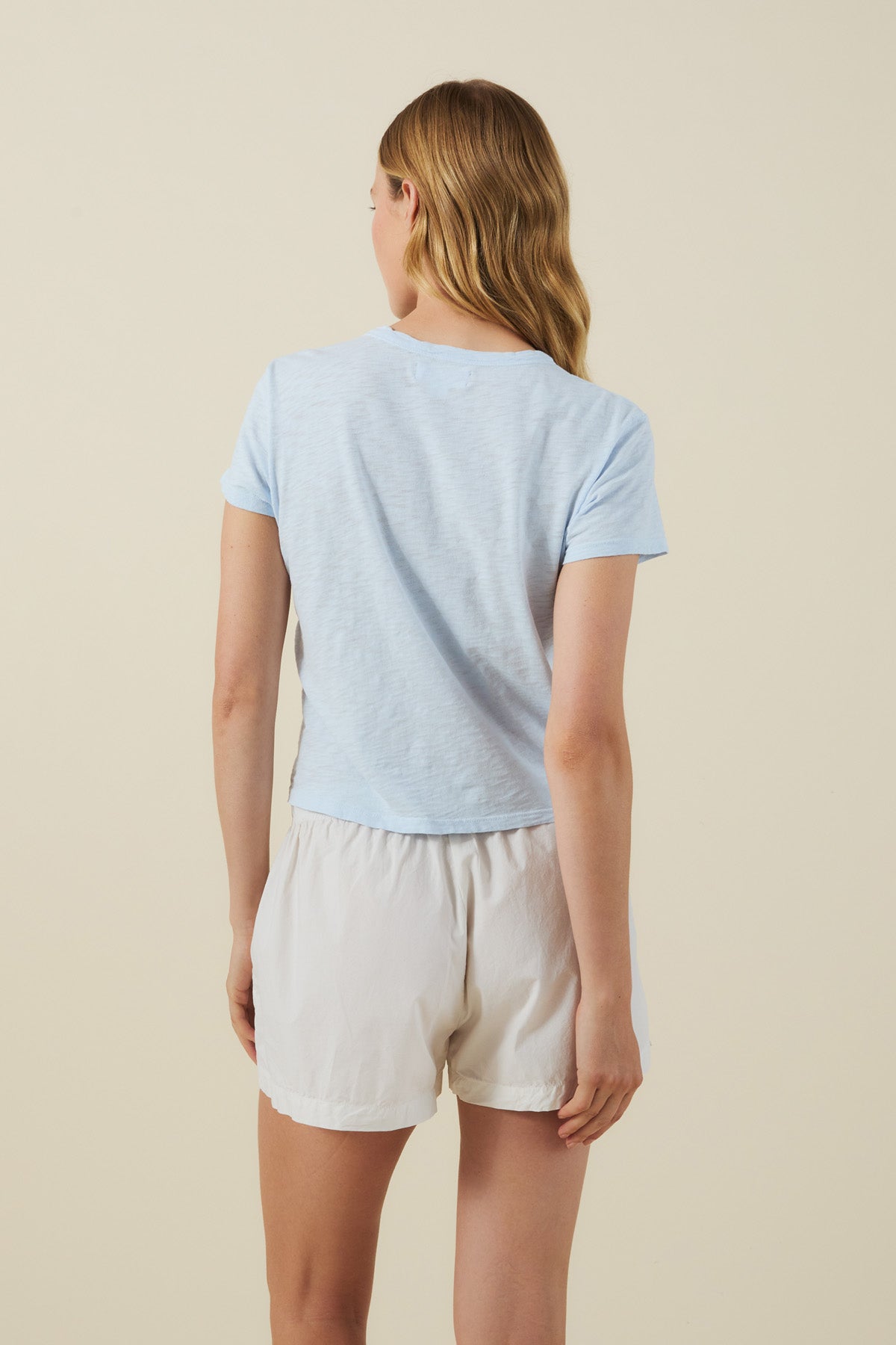 A person with long hair, seen from behind, wears the MARIKA TEE by Velvet by Graham & Spencer in light blue—a relaxed, boxy-fit short-sleeve t-shirt crafted from soft cotton slub for casual comfort—paired with white shorts against a plain beige background.