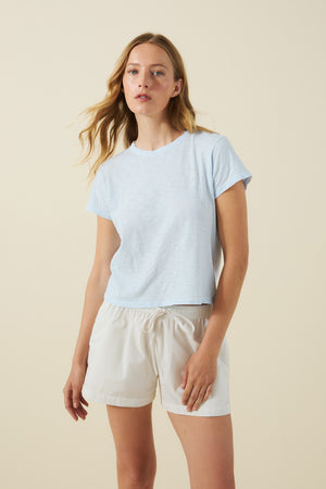 A person stands against a plain light background, wearing the Velvet by Graham & Spencer MARIKA TEE in light blue cotton slub with a relaxed boxy fit, paired with white drawstring shorts.