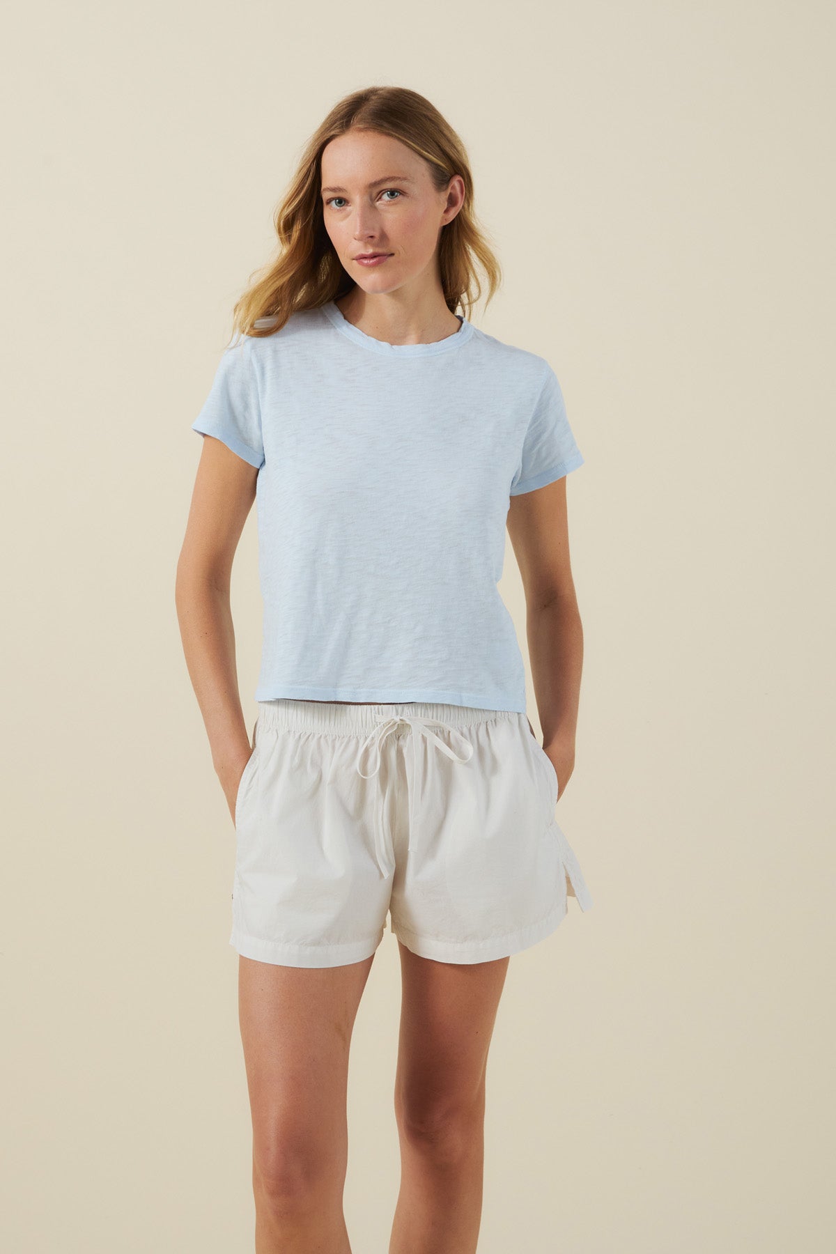 A woman stands against a neutral background, wearing the light blue MARIKA TEE by Velvet by Graham & Spencer in a relaxed boxy fit, paired with white drawstring shorts. With her hands in her pockets and a relaxed expression, the cotton slub texture of the tee lends an effortlessly casual touch to her look.-41713332158657