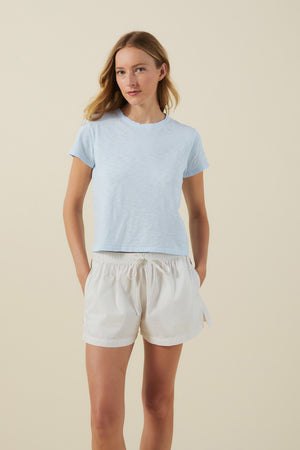 A woman stands against a neutral background, wearing the light blue MARIKA TEE by Velvet by Graham & Spencer in a relaxed boxy fit, paired with white drawstring shorts. With her hands in her pockets and a relaxed expression, the cotton slub texture of the tee lends an effortlessly casual touch to her look.