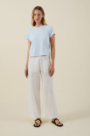 A woman stands against a plain background wearing the Velvet by Graham & Spencer MARIKA TEE in light blue—a relaxed, boxy-fit top crafted from soft cotton slub—paired with white loose-fitting pants and black sandals.
