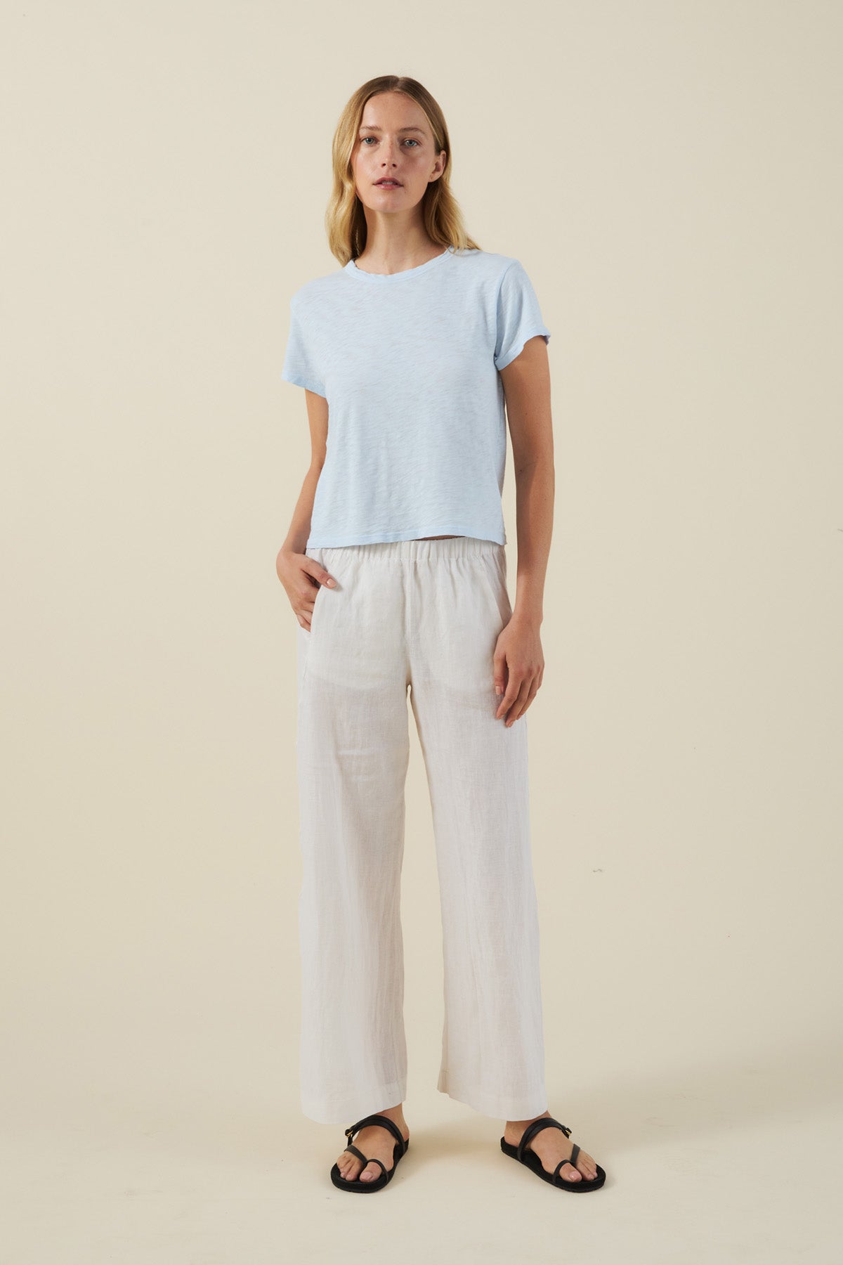 A woman stands against a beige background, wearing a light blue short-sleeve top, the LOLA LINEN PANT in white by Velvet by Graham & Spencer, and black sandals.-41779861291201