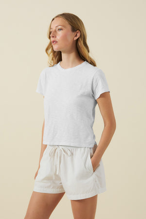 Woman stands with hands in pockets, wearing the MARIKA TEE by Velvet by Graham & Spencer in a boxy fit and white cotton slub drawstring shorts, gazing to the side against a neutral background.
