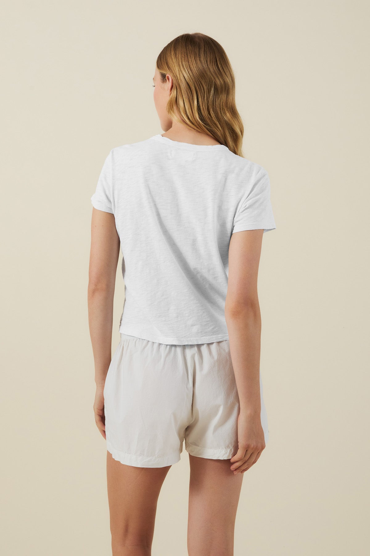 A person with long, wavy hair faces away, wearing a boxy fit MARIKA TEE by Velvet by Graham & Spencer in cotton slub with white shorts against a plain beige background.