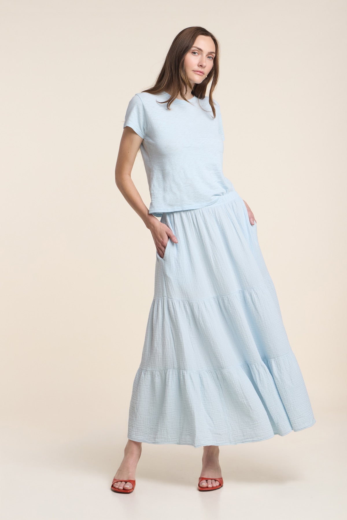 A woman wears the DANIELLE SKIRT by Velvet by Graham & Spencer—a light blue cotton gauze skirt with elastic waist and pockets—paired with a matching short-sleeve top and red sandals, set against a plain beige background.