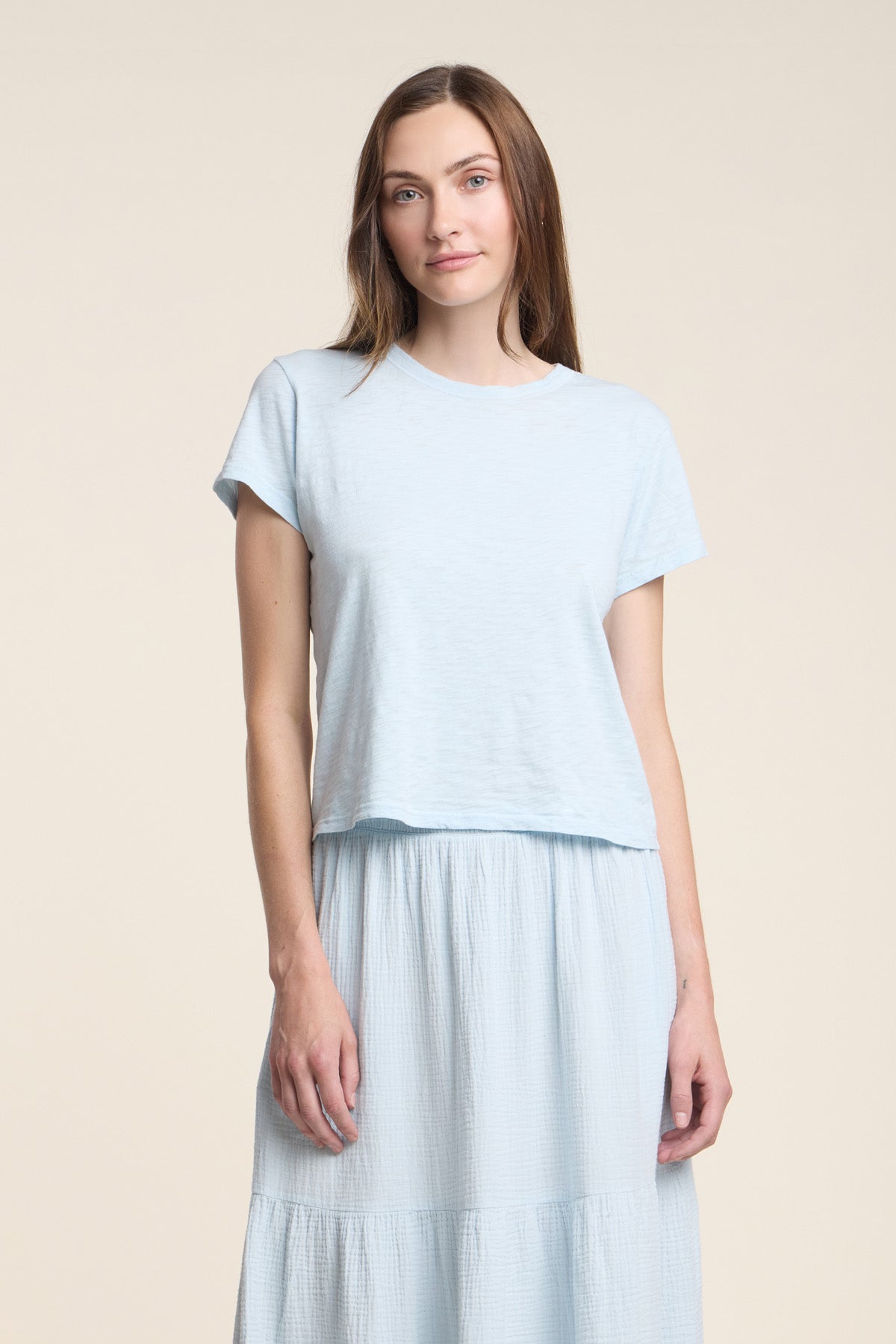 A woman faces forward, wearing the light blue cotton MARIKA TEE by Velvet by Graham & Spencer with a boxy fit and a matching long skirt, set against a plain beige background.