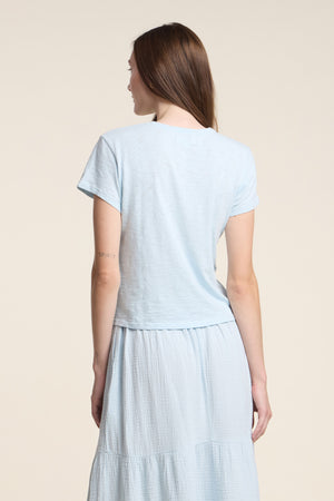 A woman with long brown hair is seen from the back wearing a light blue Velvet by Graham & Spencer MARIKA TEE in cotton slub fabric and a matching relaxed fit skirt.