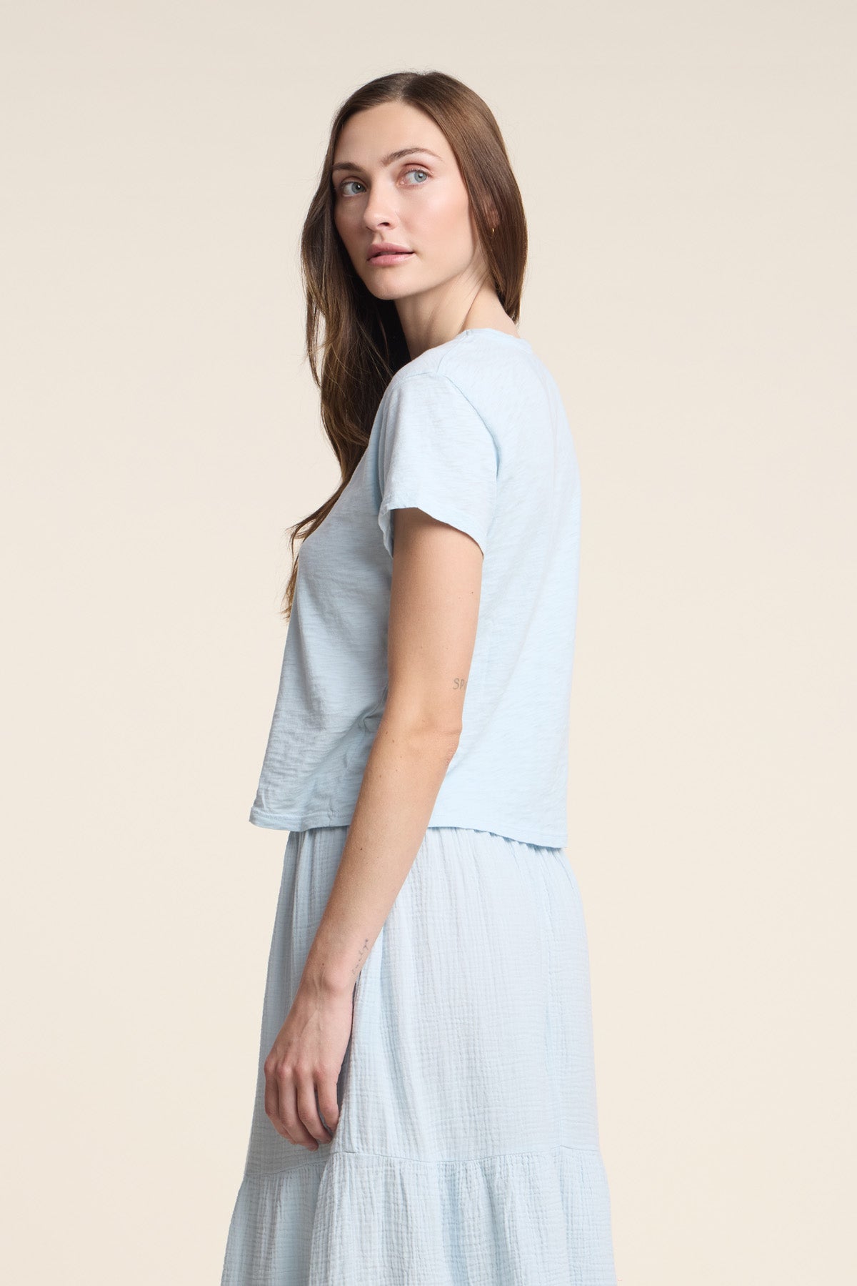 A woman with long brown hair wears a Velvet by Graham & Spencer MARIKA TEE in light blue—featuring a relaxed boxy fit in cotton slub—paired with a matching skirt, standing facing left against a neutral background.-41338479182017