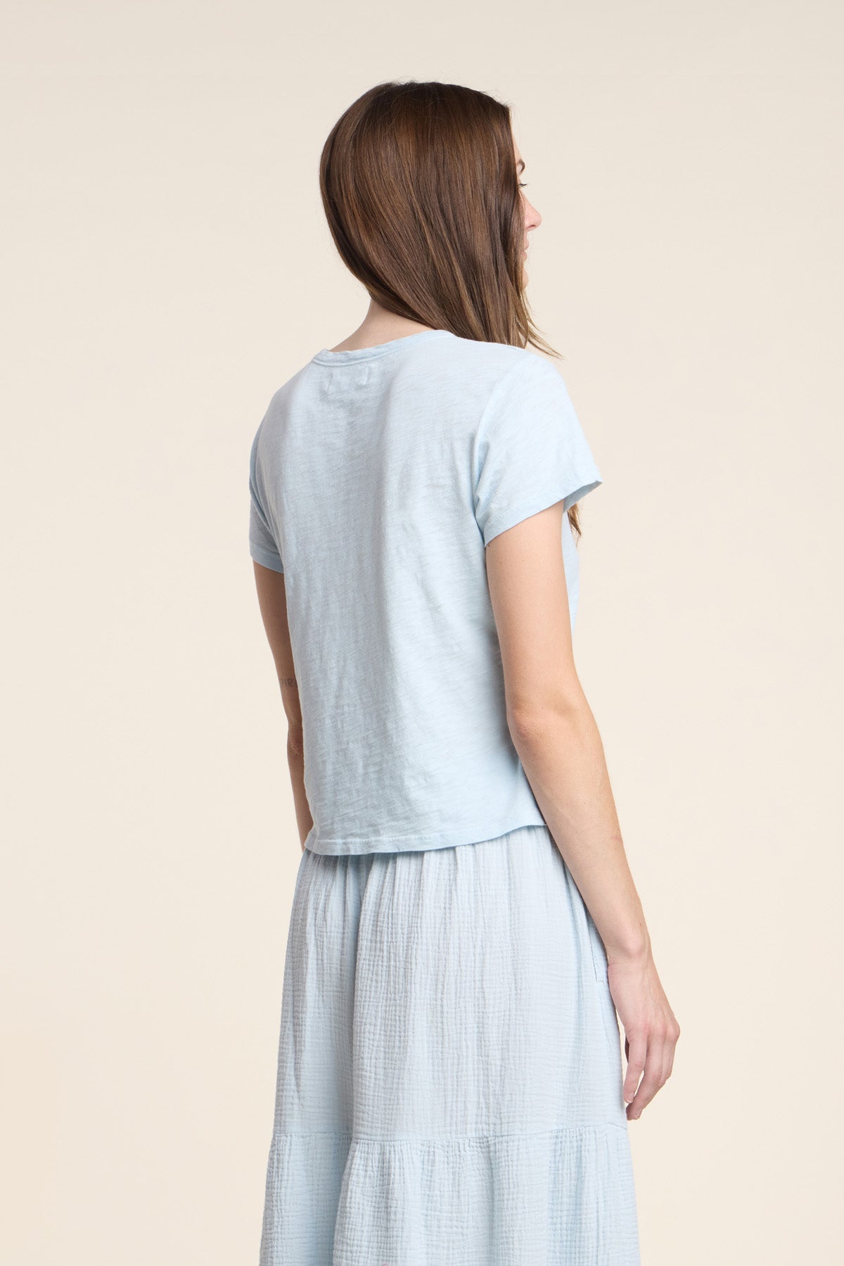 A woman stands facing away, wearing the light blue MARIKA TEE by Velvet by Graham & Spencer, crafted from cotton slub in a relaxed boxy fit, paired with a matching light blue skirt, set against a neutral beige background.