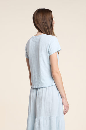 A woman stands facing away, wearing the light blue MARIKA TEE by Velvet by Graham & Spencer, crafted from cotton slub in a relaxed boxy fit, paired with a matching light blue skirt, set against a neutral beige background.