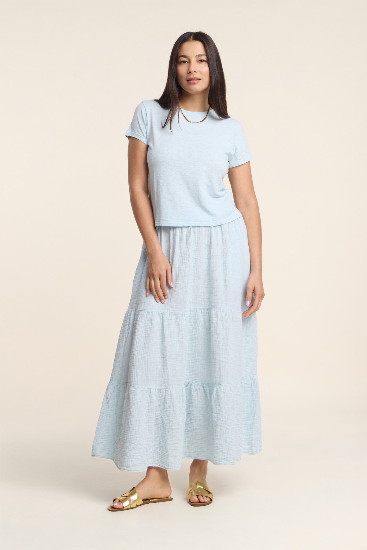 A woman faces forward in a light blue short-sleeve t-shirt and the DANIELLE SKIRT by Velvet by Graham & Spencer, a cotton gauze skirt with an elastic waist, paired with gold sandals against a plain beige background.-41338479018177