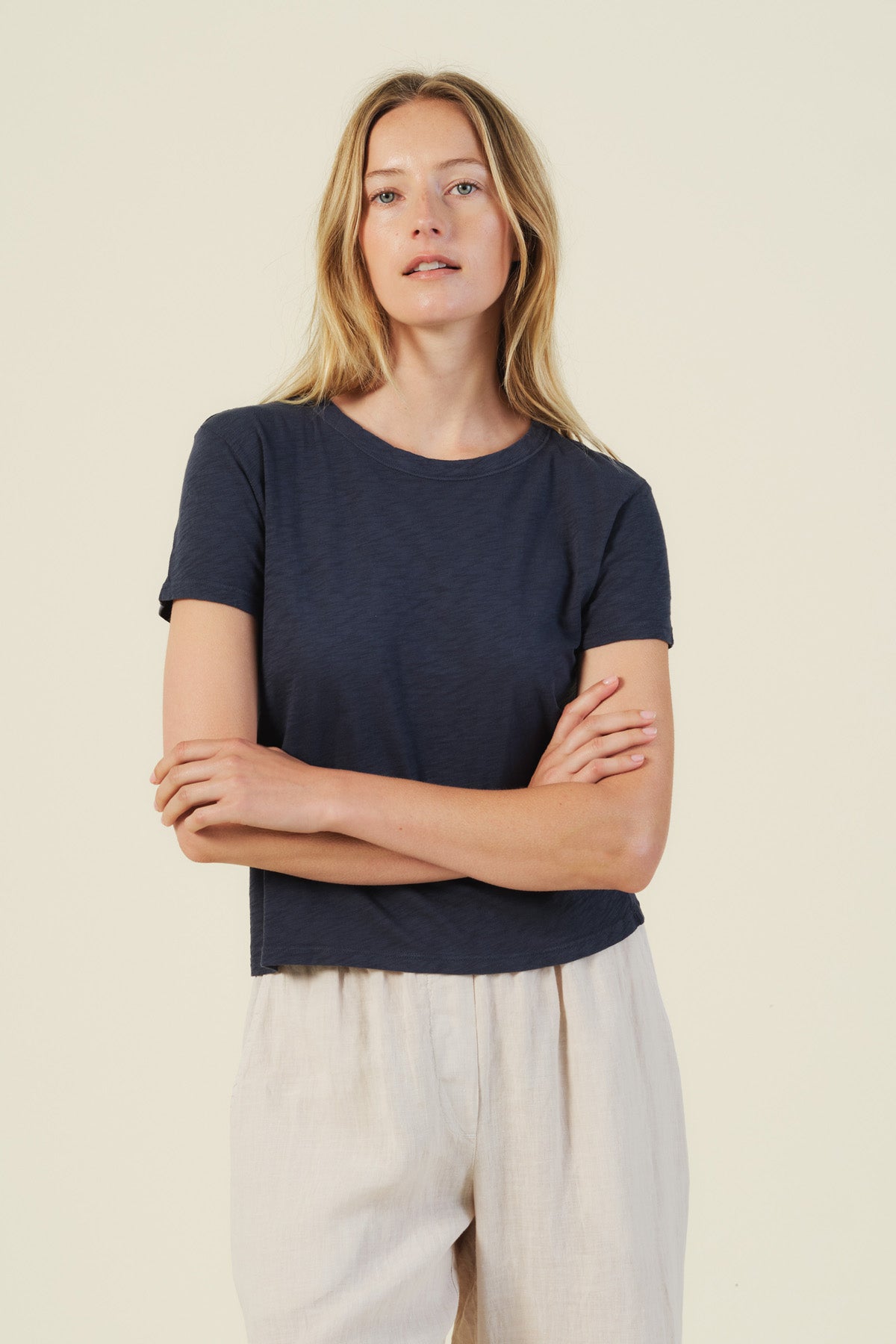 A woman with long blonde hair stands with her arms crossed, wearing the MARIKA TEE by Velvet by Graham & Spencer in navy blue cotton slub for a boxy fit, paired with light beige pants against a plain light background.
