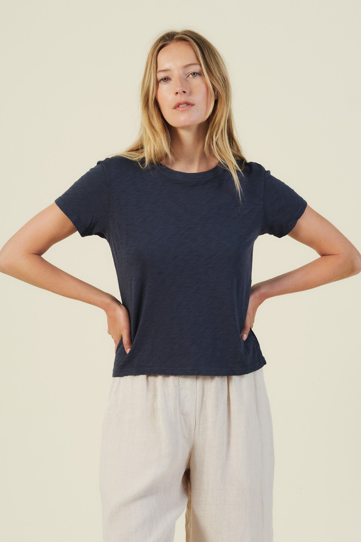 A woman stands against a plain background wearing the Velvet by Graham & Spencer MARIKA TEE in navy, featuring a boxy fit and soft cotton slub fabric, paired with light pants and her hands on her hips.-41491729973441
