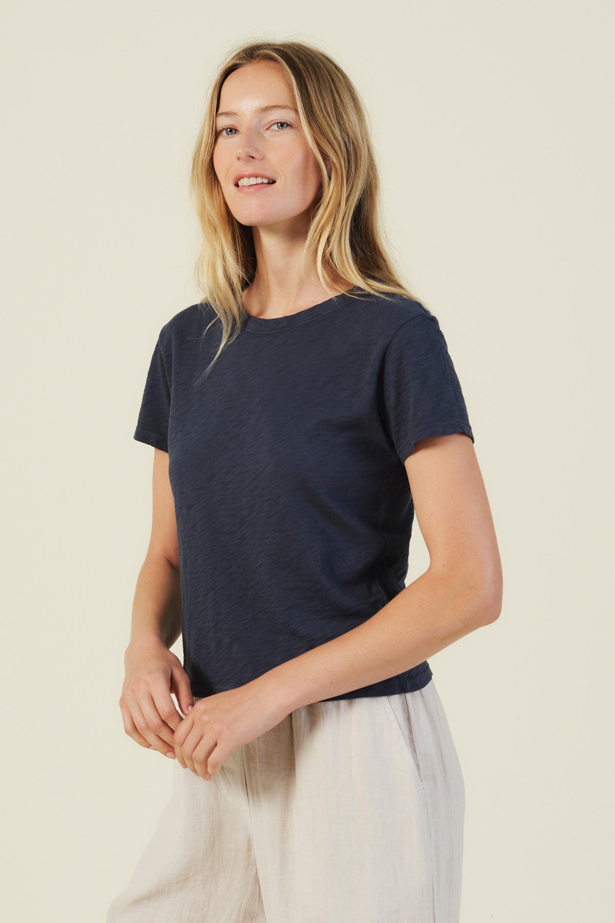 A woman with long blonde hair wears the Velvet by Graham & Spencer MARIKA TEE in navy, featuring a relaxed boxy fit and soft cotton slub, paired with light-colored pants against a plain light background.