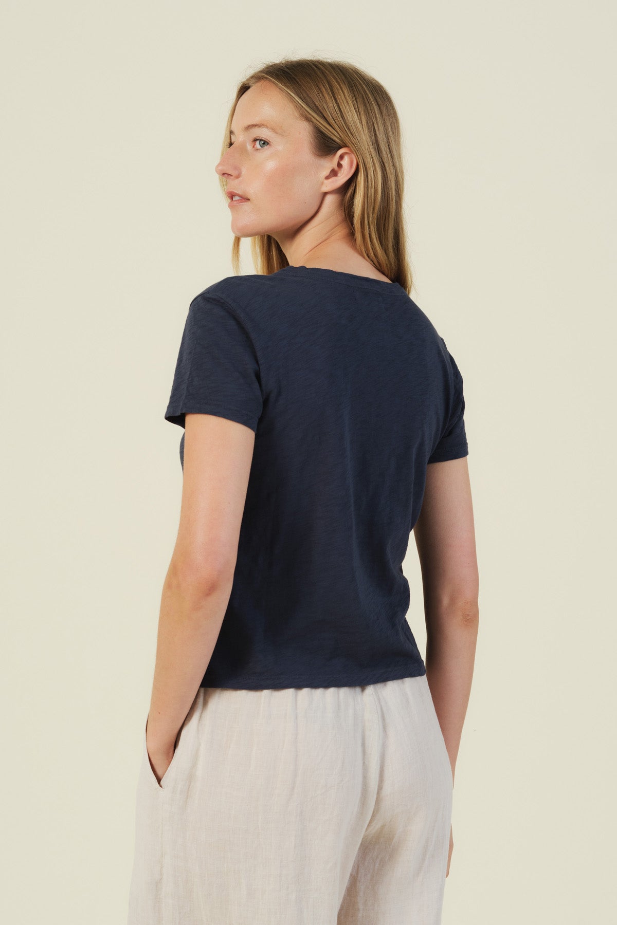 A woman stands slightly turned away, wearing a navy MARIKA TEE by Velvet by Graham & Spencer with a boxy fit and light pants against a plain background.-41491730071745