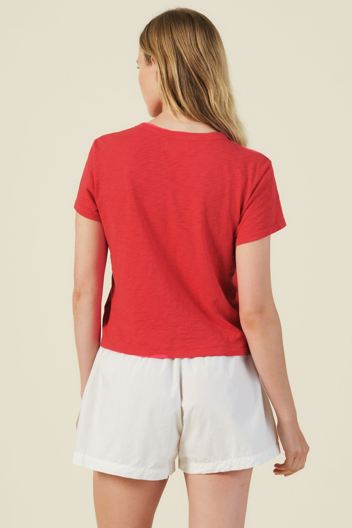 A person with long blonde hair is shown from the back, wearing a boxy fit MARIKA TEE by Velvet by Graham & Spencer and red and white shorts against a plain light background.-41387975213249