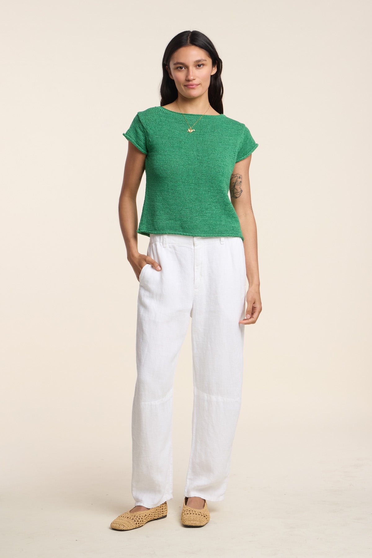 A person stands against a plain background wearing the MILOU SWEATER by Velvet by Graham & Spencer in green cotton tape yarn, paired with white loose-fitting pants and tan flat shoes, one hand in their pocket.