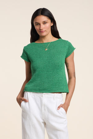 A woman wears the green MILOU SWEATER by Velvet by Graham & Spencer, paired with white pants and a gold necklace, standing with hands in pockets against a plain light background.