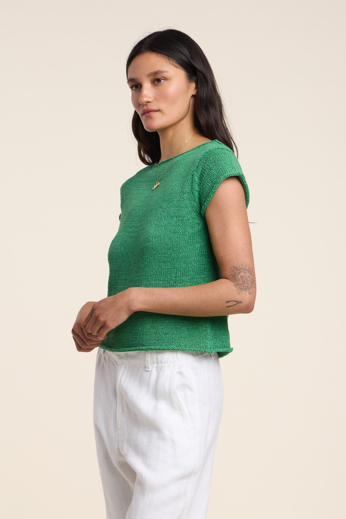 A woman with long dark hair, a forearm tattoo, and a gold necklace stands in profile wearing Velvet by Graham & Spencer's MILOU SWEATER— a green sleeveless cotton tape yarn top—paired with white pants.