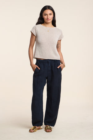 A woman faces forward with hands in her pockets, wearing the sleeveless beige MILOU SWEATER by Velvet by Graham & Spencer, loose navy pants, and gold sandals, against a plain background.
