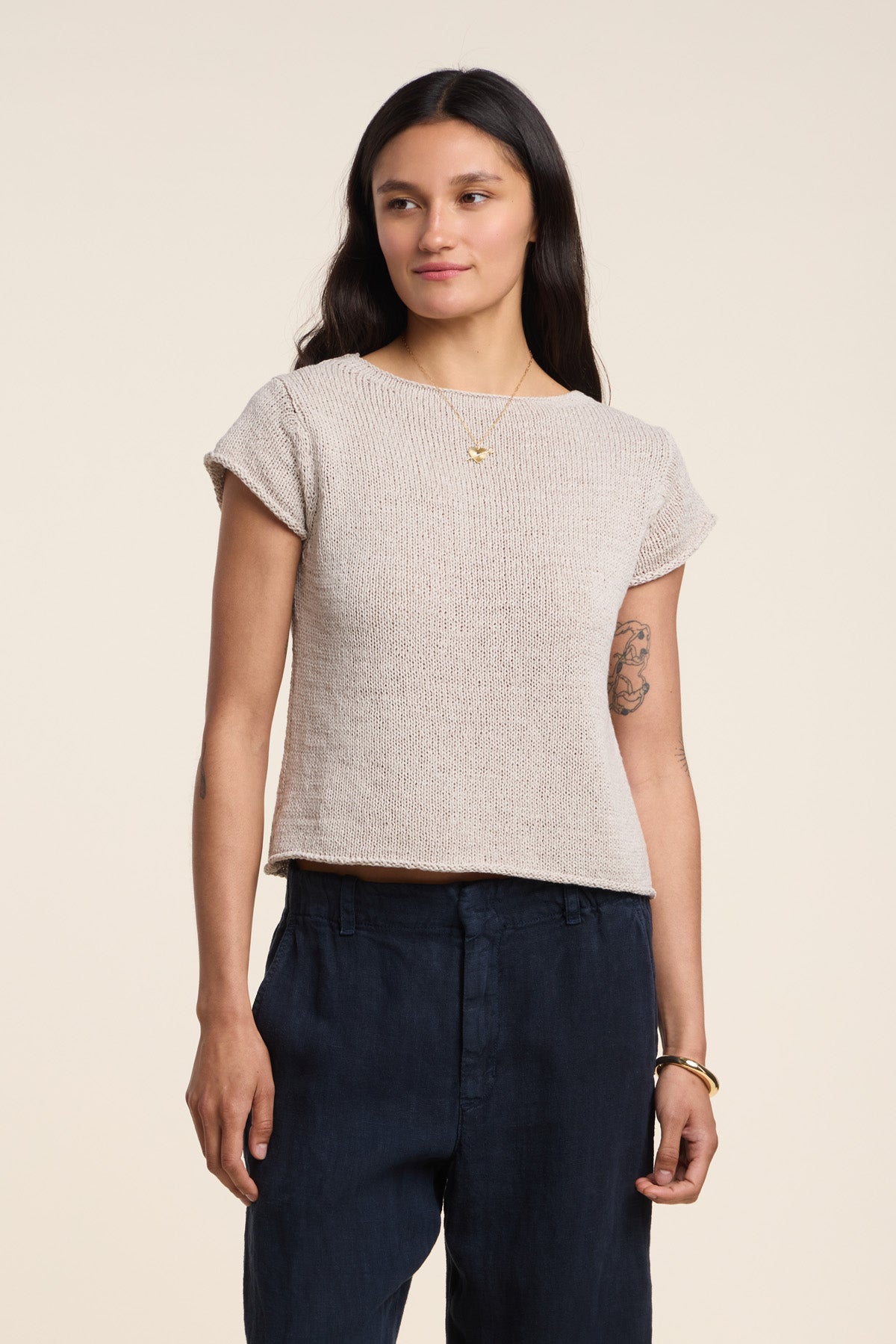 A woman with long dark hair wears the MILOU SWEATER by Velvet by Graham & Spencer, a chic beige sleeveless cotton sweater, paired with dark pants and standing against a plain light background.