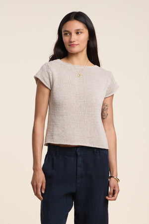 A woman with long dark hair wears the MILOU SWEATER by Velvet by Graham & Spencer, a chic beige sleeveless cotton sweater, paired with dark pants and standing against a plain light background.