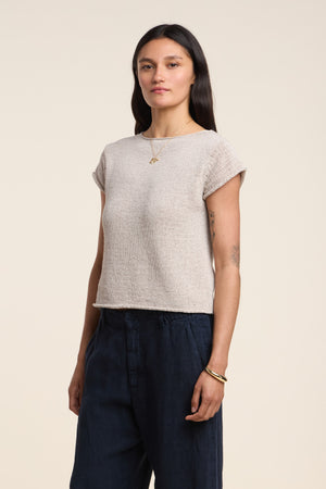 A woman stands against a plain background wearing the MILOU SWEATER by Velvet by Graham & Spencer, paired with dark pants, a necklace, bracelets, and showing tattoos on her left arm.