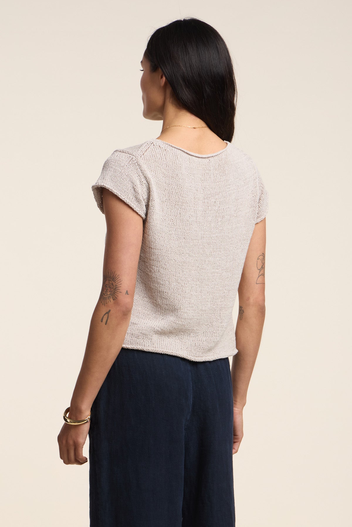 A woman stands with her back to the camera, wearing Velvet by Graham & Spencer’s MILOU SWEATER in soft cotton tape yarn and dark pants, showcasing tattoos on her arms.-41527100571841