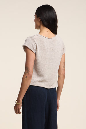 A woman stands with her back to the camera, wearing Velvet by Graham & Spencer’s MILOU SWEATER in soft cotton tape yarn and dark pants, showcasing tattoos on her arms.